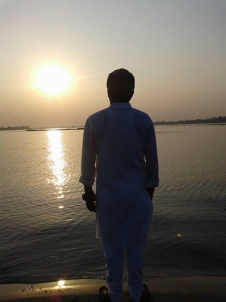 Masud Pathik seen from behind, standing at the edge of a wide river at sunset